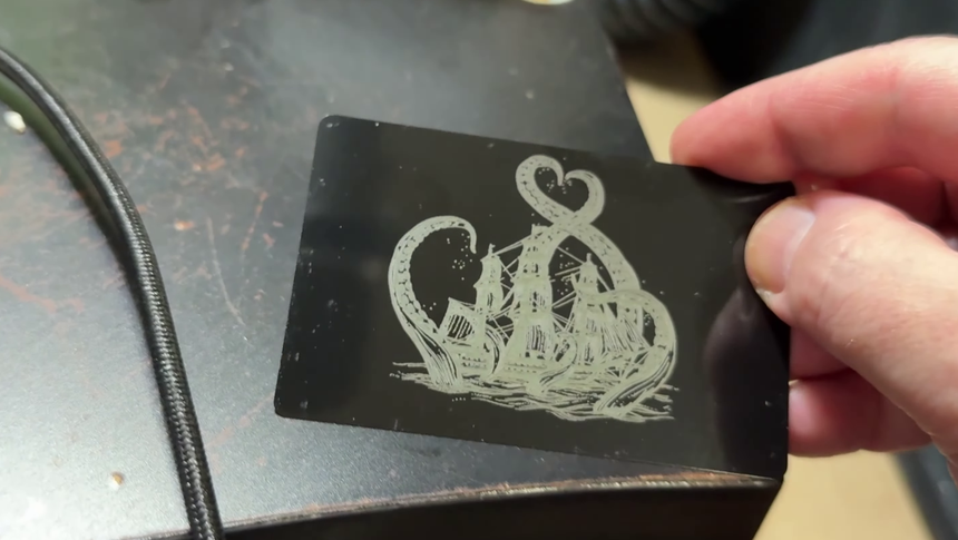 A black coated aluminum card engraved with a detailed illustration, the design showing bright silver against the black coating