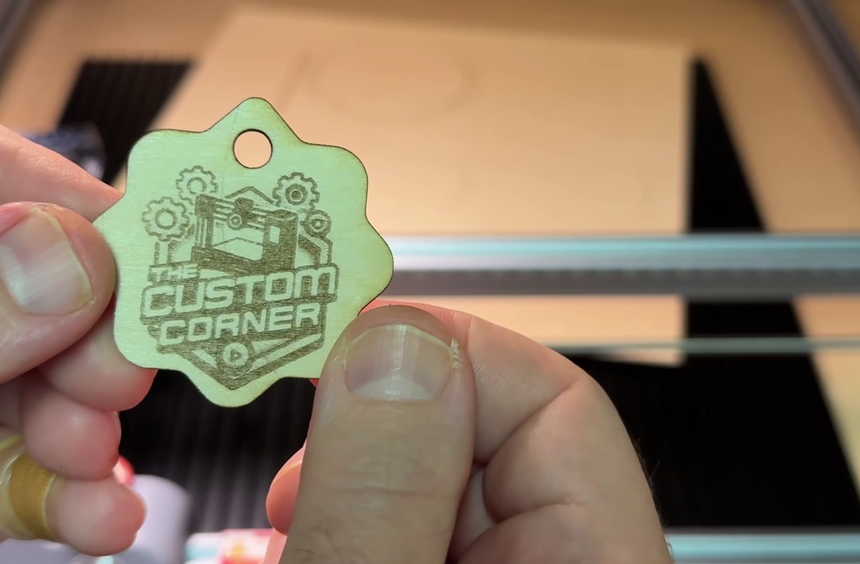 A cut and engraved balsa wood keychain with a logo, held in front of the laser