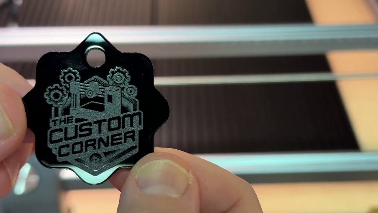 A finished black acrylic keychain with a laser engraved logo, held up in front of the laser bed